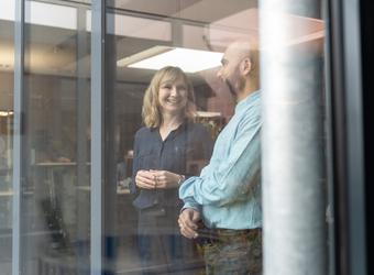 Two employees talking in a glass-walled office, viewed through large windows in a modern workplace.